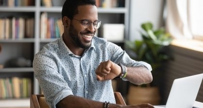 Ein Mitarbeiter arbeitet am Laptop mit einer Online Zeiterfassung und schaut auf die Uhr.