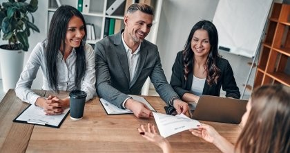 Ein Team sitzt am Tisch, hat Unterlagen und einen Laptop mit dem Bewerbungsmanagement Programm vor sich und übergibt einer Bewerberin einen Vertrag.