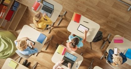 Schultische von oben mit Kindern, die an Laptops, Tablets und Smartphones arbeiten zeigen den Stand der Digitalisierung in der Schule.