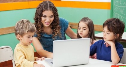 Eine Erzieherin zeigt Kindern etwas am Computer. Die Kinder verwaltet sie mit einer Kindergartensoftware.