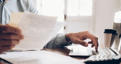 Eine Person hat Papierrechnungen in der Hand, hat einen Taschenrechner vor sich und gibt Daten in ihr Rechnungsprogramm am Laptop ein.