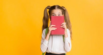 Ein Grundschulkind hält ein Buch vor das Gesicht und schaut mit großen Augen dahinter hervor.