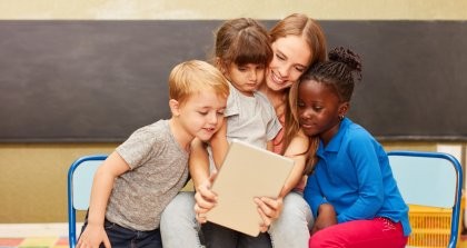 Drei Kinder und eine Erzieherin schauen auf ein Tablet vor einer Tafel auf Stühlen